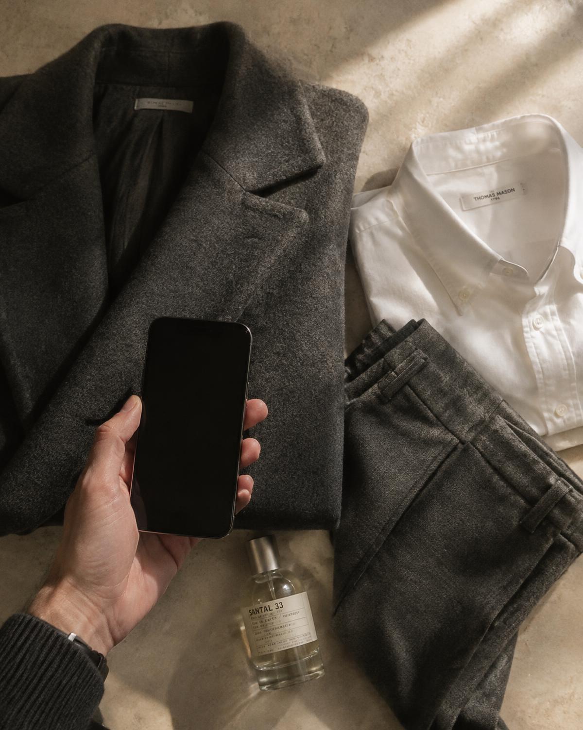 A hand holding a phone over a laid-out charcoal overcoat, flannel trousers and white shirt